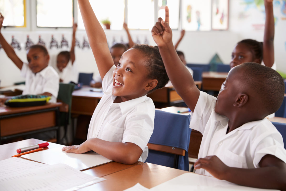 A group of children sitting at desks raising their hands.